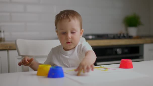 Lindo niño sentado detrás de la mesa y dibujando con los dedos cubiertos de pintura colorufl. Concepto de educación infantil, desarrollo creativo y arte en el hogar — Vídeo de stock