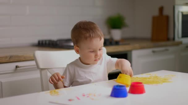 Lindo niño sentado detrás de la mesa y dibujando con los dedos cubiertos de pintura colorufl. Concepto de educación infantil, desarrollo creativo y arte en el hogar — Vídeo de stock