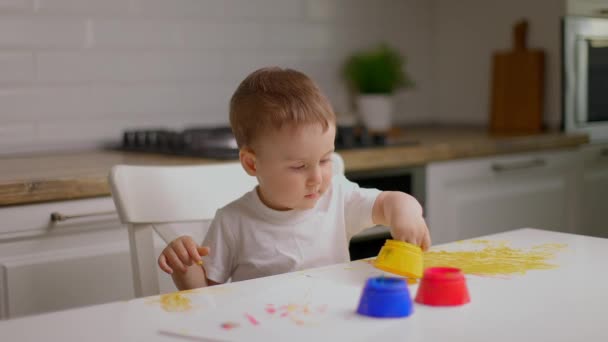 Słodki chłopczyk siedzi za stołem i rysuje palcami pokrytymi farbą kolorową. Koncepcja edukacji dzieci, rozwoju kreatywności i sztuki w domu — Wideo stockowe
