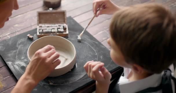 Мальчик и мать вместе украшают глиняную чашу буквами. Мастер-класс в мастерской по керамике. Вид сверху. Медленное движение — стоковое видео