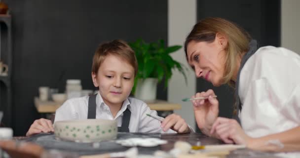 Moeder pottenbakker helpt zijn zoon met het maken van patroon op de schaal in pottenbakkerij studio. Masterclass in een pottenbakkerij. Langzame beweging — Stockvideo