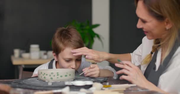 Moeder pottenbakker helpt zijn zoon met het maken van patroon op de schaal in pottenbakkerij studio. Moeder streelt liefdevol zijn zoon op het hoofd. Langzame beweging — Stockvideo