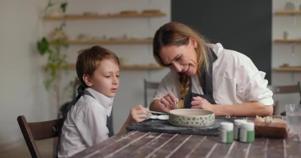 Vrouwelijke blanke pottenbakker helpt zijn zoon met het maken van een patroon op de kleikom. Langzame beweging — Stockvideo