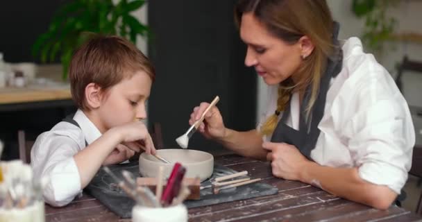 Anne ve oğlu çömlek atölyesinde, atölyede birlikte kilden bir kase çiziyorlar. Kadın çömlekçi çocuğa toprak eşyalar üzerinde resim yapmayı öğretiyor. Yavaş çekim — Stok video