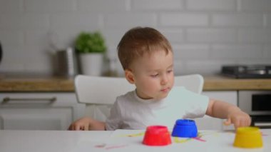 Bebek çocuk masanın arkasında oturuyor ve evde boyayla boyanmış parmaklarıyla resim çiziyor.