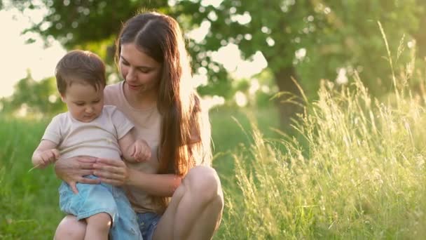 Mãe feliz com o seu filho sorridente no jardim. Jovem mãe e seu bebê bonito brincando em flores. O bebé cheira a flor. Movimento lento — Vídeo de Stock