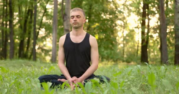 Jongeman die op het gazon zit en mediteert in het stadspark. Mannen die op gras zitten in lotus poseren mediterend buiten bij zonsopgang. Langzame beweging — Stockvideo