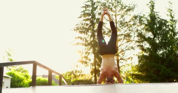 Uomo sportivo in piedi a yoga posa all'aperto la mattina.Guy bilanciamento sulla testa e lentamente muovendo le gambe verso il basso. Bei alberi come sfondo. — Video Stock