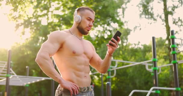 Człowiek przygotowuje się do treningu na boisku sportowym.Sportowiec nosi słuchawki i trzyma telefon — Wideo stockowe