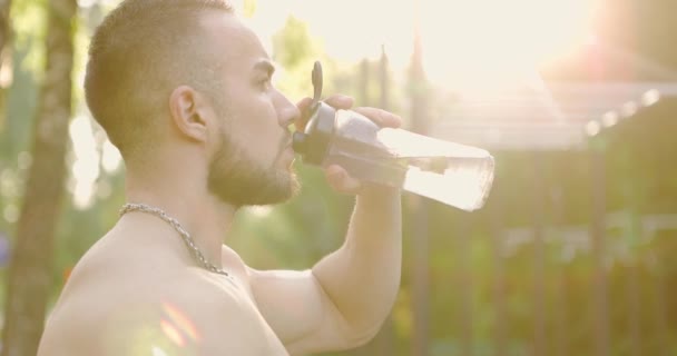 Muskelsportler entspannt sich nach dem Training auf Sportplatz am Morgen. Mann trinkt Wasser feuchtigkeitsspendend — Stockvideo