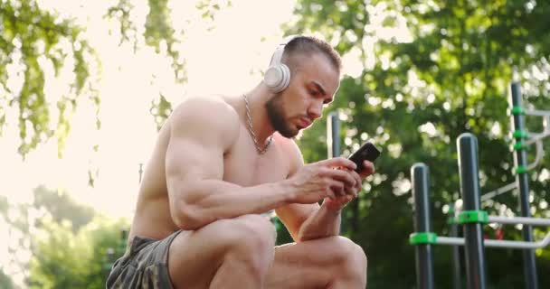 Uomo muscoloso seduto sul campo sportivo nel parco della città e controllare il telefono mentre listerning alla musica — Video Stock