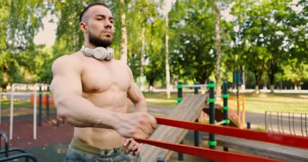 Mann macht Übungen mit Gummiband im Stadtpark.Bodybuilder trainiert seine Arm- und Rückenmuskulatur. — Stockvideo