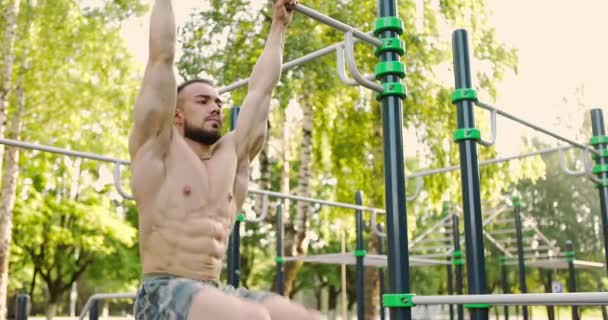 Νεαρός γενειοφόρος άνδρας με γυμνό κορμό κάνει ανύψωση ποδιών σε οριζόντια ράβδο σε αθλητικό έδαφος — Αρχείο Βίντεο