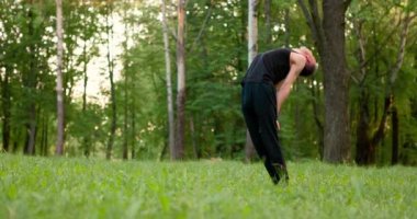 Siyah elbiseli genç adamlar güneşli yaz gününde parkta yoga ve meditasyon yapıyorlar. Sırtı bükük duruyor. Yavaş çekim