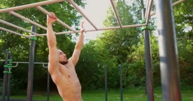 Parktaki spor sahasında kaslı, üstsüz bir adamın maymun tezgahında sallanışının yan görüntüsü. Fitness sporu açık havada brachiation ladderin üzerinde taşınıyor. Spor ve sağlıklı yaşam tarzı kavramı