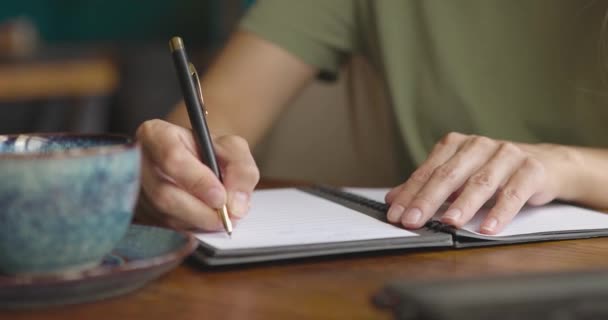 テーブルの上にコーヒーカップとラップトップで紙のノートブックに書いている女性の手の閉じる — ストック動画