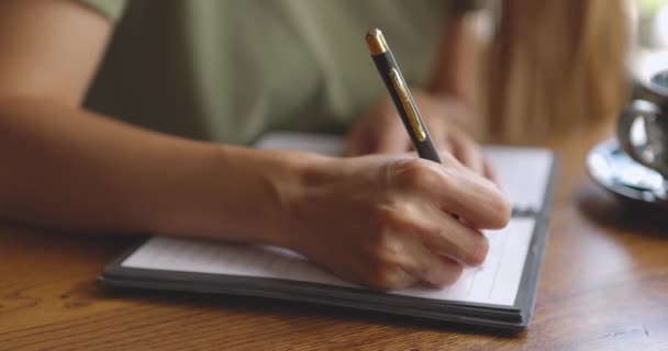 Primer plano de las manos de las mujeres que escriben en un cuaderno blanco en blanco con taza en la mesa de madera — Vídeo de stock