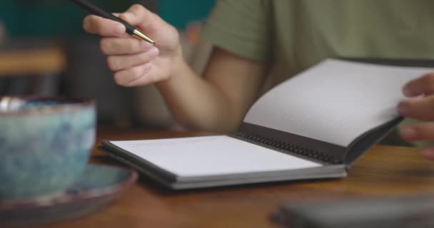 Primer plano de las manos femeninas abriendo cuaderno de papel y tomando notas.Mujer trabajando en un acogedor café — Vídeo de stock