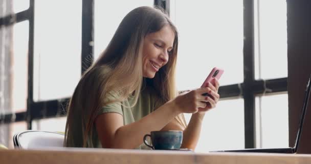 Jovem com cabelos longos conversando usando smartphone, rindo enquanto toma uma xícara de café no café — Vídeo de Stock