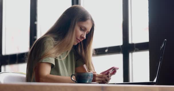 작은 레스토랑에서 노트북을 가지고 일하는 여성 커피를 들고 앉아서 전화로 타이핑하는 여성 — 비디오