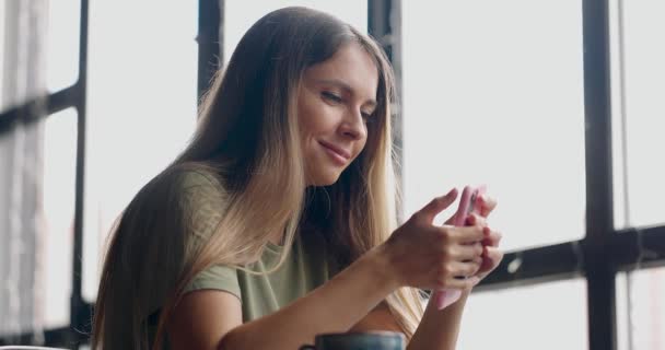 Kvinna som äter lunch i caféet, dricker kaffe, ler, håller rosa smartphone och skickar meddelande — Stockvideo