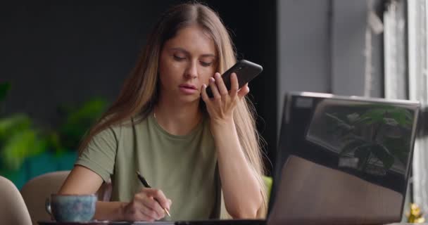Žena sedí v kavárně a drží pero a dělá si poznámky do deníku, směje se, mluví na reproduktoru přes telefon — Stock video