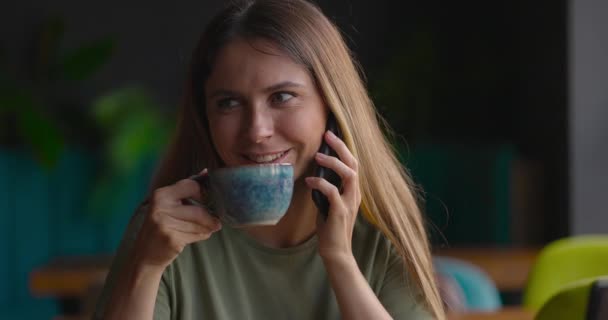 Nahaufnahme einer attraktiven Frau mit langen Haaren, die Kaffee trinkt und glücklich im Café telefoniert — Stockvideo