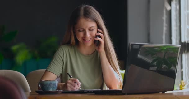 Csinos nő jegyzetel egy jegyzetfüzetbe, miközben telefonál és mosolyog a kávézóban. — Stock videók