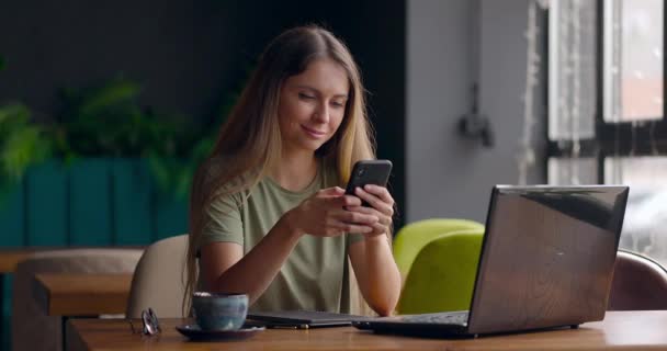O femeie drăguță trimite mesaje folosind telefonul mobil și zâmbește în timp ce lucrează cu laptopul în cafenea — Videoclip de stoc