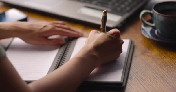 Primer plano de la escritura de la mano de las mujeres en papel notebook.Cup de té negro, ordenador portátil y teléfono en la mesa — Vídeos de Stock