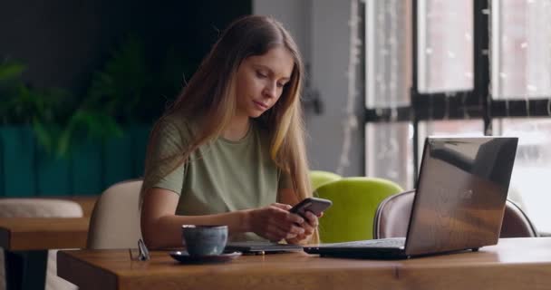 Žena sedí v kavárně a psaní na telefonu Freelancer pracuje on-line pomocí notebooku a pití kávy — Stock video