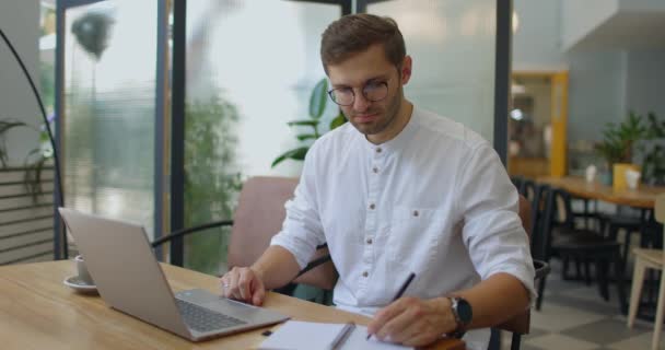 Bilgisayarda video görüşmesi yapan ve küçük bir kafede otururken not alan bir adam. — Stok video