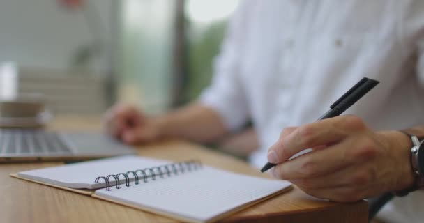 Manos de hombre escribiendo notas en cuaderno con bolígrafo negro sentado con el ordenador portátil en la mesa de café de madera — Vídeo de stock