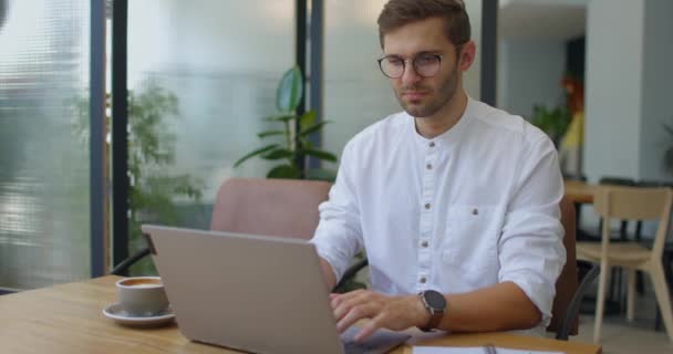 Hombre joven en camisa sentado en la cafetería, trabajando en línea, sitios web de desplazamiento y charlando con el ordenador portátil — Vídeos de Stock