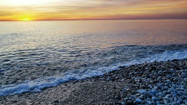 Deniz dalgaları yavaşça kıyıya yuvarlıyor, çakıl taşlarını yuvarlıyor ve geri geliyor. Güneşli bir gün batımı gökyüzünü parlak renklerle boyar.
