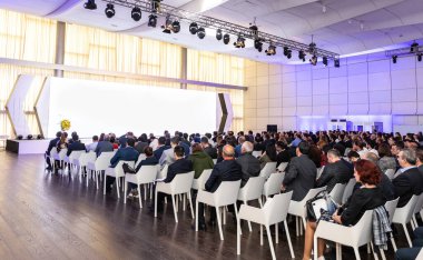 Konferans salonundaki seyirciler, insanlar ekrana bakıyor.