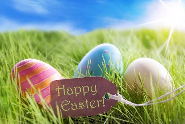 Drie kleurrijke Pasen eieren op zonnige groen gras met Label vrolijk Pasen — Stockfoto