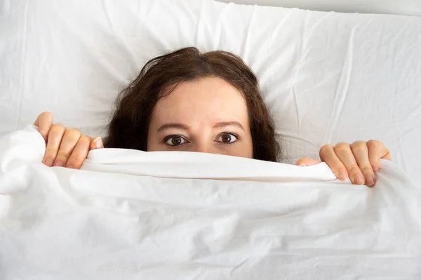 Young scared woman hiding under blanket while lying in bed at home