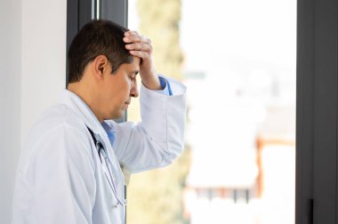 Genç bir erkek doktorun, hastanenin penceresinde dikilirken stresli bir şekilde bakması.