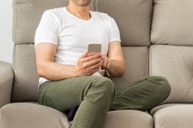 Oturma odasındaki kanepede oturan telefona bakan bir adamın bacakları. Yazın evin içinde.