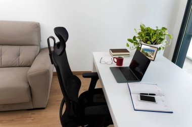 Gündüz vakti boş bir ofisteki masada dizüstü bilgisayara odaklanmış bir defter ve teknolojinin kesilmiş görüntüsü.