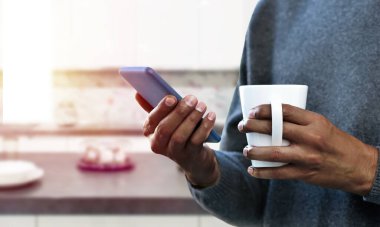 Elleri kahve bardağını tutan bir adam, telefonu mutfağın içinde tutuyor.