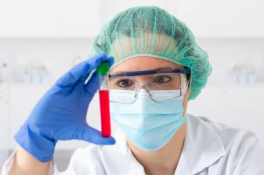 Koruyucu yüz maskesi takan ve gözlük takan genç bir kadın laboratuarda test tüpünü inceleyerek gözlere odaklanıyor. 