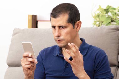 Evdeki oturma odasındaki koltukta oturan akıllı telefona bakan kızgın adam.