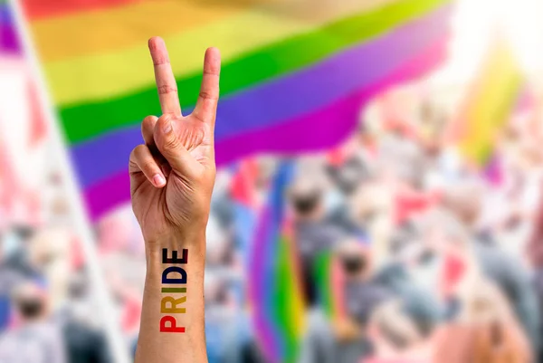 Supporting hand makes peace sign in front of a rainbow flag flying on the sidelines of a summer gay pride parade with copy space