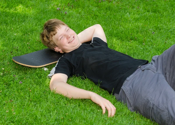 Ragazzo ridente rilassarsi sullo skateboard in erba parco — Foto Stock