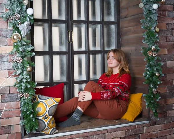 Ein blondes Mädchen im roten Pullover sitzt auf der Fensterbank. — Stockfoto