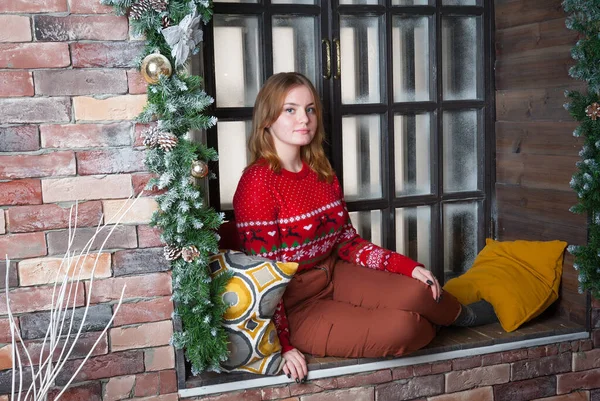 En blond tjej i röd tröja sitter på fönsterbrädan.. — Stockfoto