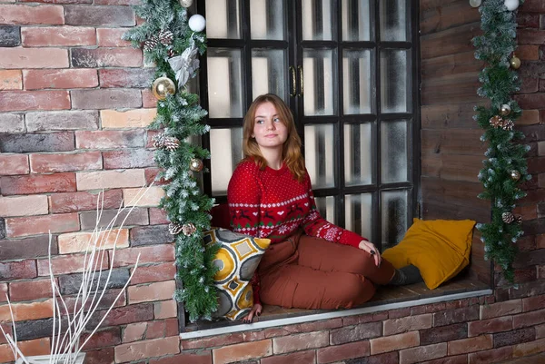 Ein blondes Mädchen im roten Pullover sitzt auf der Fensterbank. — Stockfoto
