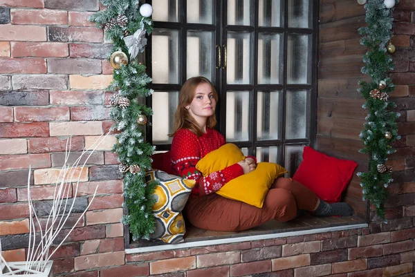 A blonde girl in a red sweater sits on the windowsill. — Stock Photo, Image